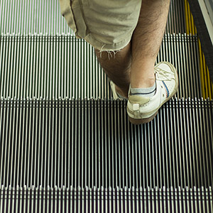 A person's feet in white sneakers and shorts, standing on a moving escalator.  